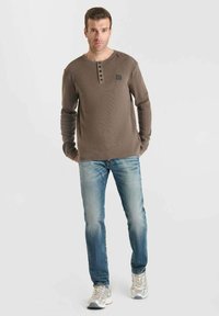 Un hombre está de pie de frente, vistiendo una camisa henley de manga larga marrón, jeans azules descoloridos y zapatillas de color claro contra un fondo liso.