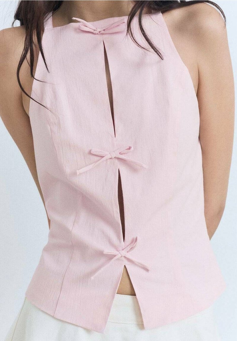 Femme portant un haut sans manches rose clair avec trois fentes verticales à l'avant nouées avec des nœuds en tissu assorti, associé à un pantalon blanc.
