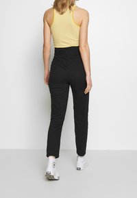 Černé kalhoty s vysokým pasem, přiléhavý střih, hladká textura, doplněné žlutým pruhovaným topem na ramínka a bílými teniskami. Záběr zezadu.