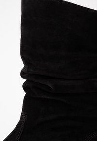 Bottes chevilles en suède noir avec un design froncé, présentant une texture lisse et des détails de coutures subtiles le long des bords.