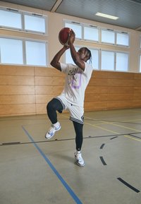 Basketbola spēlētājs baltā T-kreklā un rakstainās šortās, ar melnām legingām un zilām un baltām sporta apaviem, izpilda metienu uz grozu telpās.