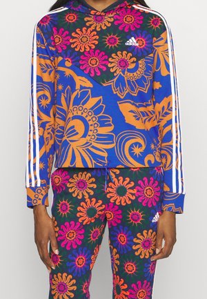 Persoon draagt een blauwe Adidas-hoodie en broek met opvallende oranje, roze en paarse bloemen- en zonnestraalpatronen en witte Adidas-strepen op mouwen en benen.