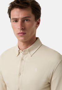 Beige overhemd met knopen en een zachte textuur, voorzien van een kraag en een klein geborduurd "B"-logo aan de linkerkant.