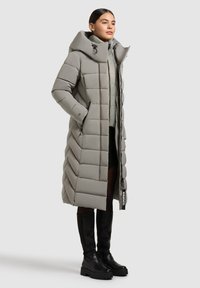 Lang grijze pufferjas met grote capuchon, chevron-gestikte patroon, twee zijzakken en een merklabel, gecombineerd met kniehoge zwarte laarzen.