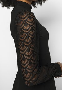 Haut col roulé noir avec manches longues en dentelle transparente présentant des motifs géométriques sur une personne aux cheveux bouclés.