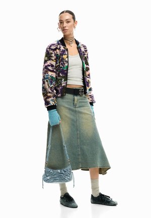 Femeie purtând o jachetă mov cu model, maiou gri, fustă midi din denim, mănuși albastre fără degete, ținând o geantă albastră și încălțări negre cu șosete gri.