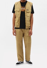 Man med beige arbetsväst och cargobyxor, svart grafisk t-shirt, svartvita sneakers, står mot en enfärgad bakgrund.