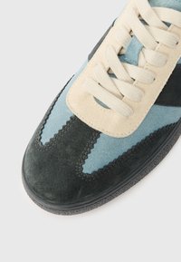 Melna, zila un bēša zamša sporta apavi ar bēšām auklām, parādot sīkās šuves un teksturētu gumijas zoli uz balta fona.