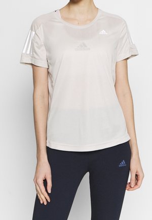 Kvinde iført lys beige Adidas kortærmet sportstrøje og mørkeblå Adidas leggings, stående mod en ensfarvet baggrund.