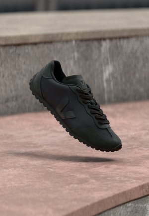 Fekete sportcipő velúr és szövet anyagból, fekete fűzőkkel és texturált talppal, kőfelület felett felfüggesztve a szabadban.