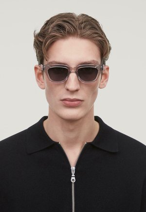 Clear rectangular sunglasses with dark lenses and a transparent frame, worn by a model in a black collared zip-up shirt.