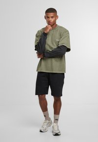 Noor mees seisab mõtlikult, käsi lõual, seljas roheliste ja mustade kihtidega särke, mustad lühikesed püksid, beežid sokid ja valged spordijalatsid.