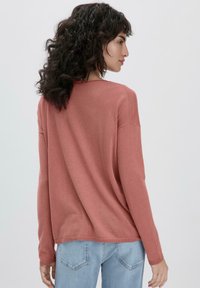 Rustfarbenes Sweatshirt mit langen Ärmeln, lockerer Passform und rundem Ausschnitt. Das Material wirkt weich und hat eine glatte Textur. Kombiniert mit hellblauen Jeans.