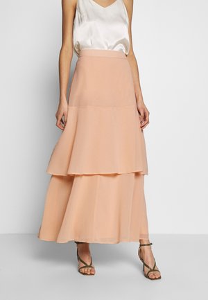 Vrouw die een perzikgekleurde gelaagde maxi-rok draagt, een witte satijnen mouwloze top en olijfgroene gespen sandalen met hoge hak, staand tegen een eenvoudige achtergrond.