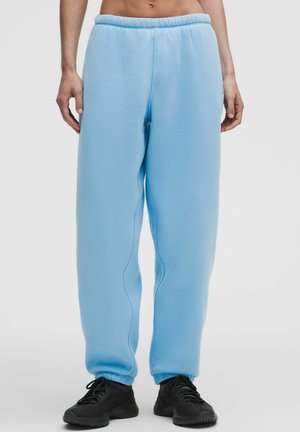 Hellblaue Fleece-Sweathosen mit elastischem Bund und Bündchen, getragen mit schwarzen Sportschuhen an einer stehenden Person vor weißem Hintergrund.