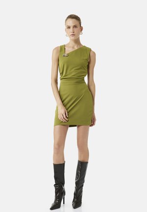 Vrouw draagt een mouwloze asymmetrische olijfgroene jurk met decoratieve speld, gecombineerd met zwarte kniehoge laarzen met hakken, staand tegen een witte achtergrond.