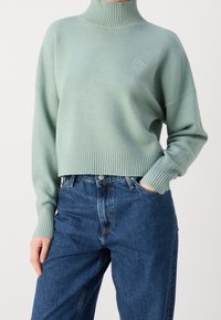 Mint green turtleneck sweater with ribbed cuffs and hem, featuring a small circular logo on the chest, paired with high-waisted blue jeans.