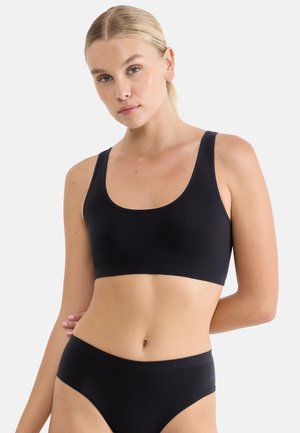 Eine Frau mit hellem Teint und blondem Haar trägt einen schwarzen Soft-BH und passende schwarze Unterwäsche vor einem schlichten hellgrauen Hintergrund.