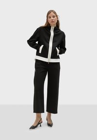 Crna zip-up dukserica s bijelim akcentima i džepovima, u kombinaciji s crnim širokim hlačama i crnim sandalama na remen. Glatka tekstura tkanine.