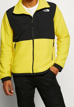 Mężczyzna noszący żółtą i czarną kurtkę polarową z kieszeniami na suwak i logo The North Face na piersi, w połączeniu z czarnymi spodniami.