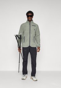 Giacca in pile di colore verde chiaro con zip completa, tasche laterali e dettagli scuri. I pantaloni neri e i bastoni da trekking completano l'outfit.
