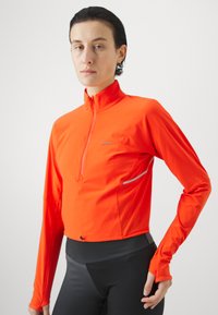 Lys orange kvart-glidelås langermet topp laget av lett stoff, med reflekterende detaljer og cropped design.