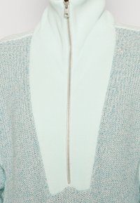 Hellblauer Strickpullover mit hohem Kragen und Reißverschluss, mit strukturierter Oberfläche und einem kontrastierenden glatten Einsatz auf der Vorderseite.