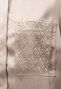 Jasnobeżowa bluzka o fakturze satyny, z dekoracyjną kieszenią w złoty wzór diamentowy. Płaskie szwy i gładki materiał.