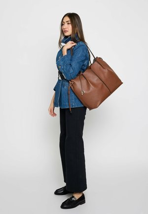 Frau trägt eine blaue Jeansjacke, schwarze Hose, schwarze Schuhe und trägt eine große braune Ledertragetasche über der Schulter vor einem schlichten Hintergrund.