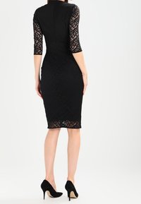 Musta cocktaildress, jossa on istuva pitsibuusti ja kolme neljänneskautta hihat. Hame on sileä ja polven pituinen, yhdistettynä mustiin korkokenkiin.