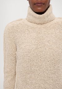 Femme portant un pull à col roulé texturé beige, visible des épaules au menton sur un fond uni.