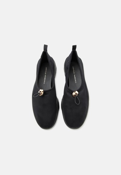 Μαύρα παπούτσια slip-on με υφασμάτινη υφή, ελαστικό κορδόνι και χρυσό κλείσιμο τύπου toggle, καθώς και ταινίες για το πίσω μέρος της φτέρνας, εμφανιζόμενα από πάνω σε λευκό φόντο.