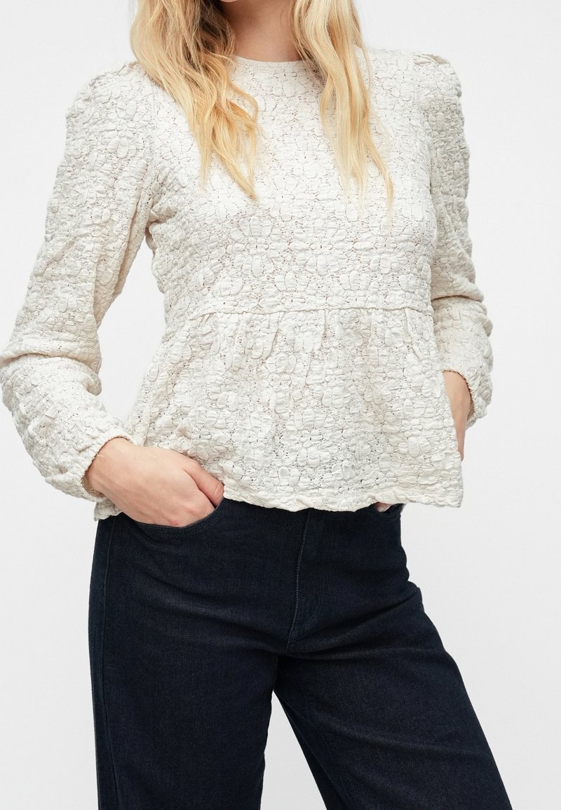 Femme portant un chemisier blanc texturé à motifs floraux, manches longues et épaules bouffantes, et un pantalon noir, les mains dans les poches.