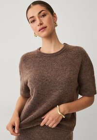Brun strikket sweater med korte ærmer og rund halsudskæring. Har en blød tekstur. Guldarmbånd på hånden. Enkel stribet baggrund.