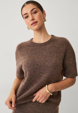 Brown knit sweater with short sleeves and a round neckline. Features soft texture. Gold bracelet on hand. Simple striped background.