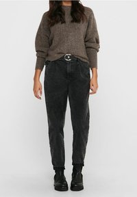 Mujer vestida con un suéter de punto marrón, jeans negros de cintura alta con un cinturón de doble anilla y botas negras por el tobillo, de pie frente a un fondo liso.