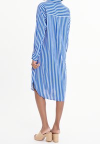 Žena v dlouhých rukávech šatů s vertikálními modro-bílými pruhy, s manžetami na knoflíky a béžovými pletenými lodičkami na podpatku, stojící na bílém pozadí.