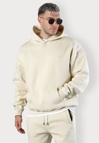 Beige oversized hoodie with a front pocket and drawstring hood, made from soft material. Model wears sunglasses and matching beige joggers.