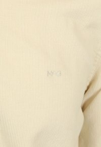 Cremefarbener, gerippter Stoff mit einer subtilen, glänzenden Textur; verfügt über ein silbernes, gesticktes "M◊G"-Logo auf der Vorderseite.