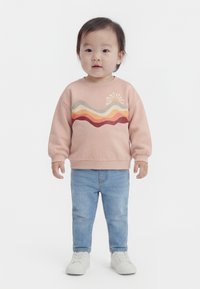 Tout-petit debout, portant un sweat-shirt pêche avec des rayures ondulées et un motif de soleil, un jean bleu clair et des baskets blanches sur fond blanc.