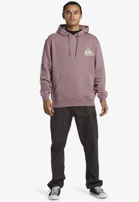 Light purple hoodie with a front pocket, drawstring hood, and logo on the chest. Paired with dark cargo pants and black sneakers.