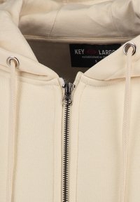 Beige zip-up hoodie met een gladde textuur, voorzien van een zilveren rits, een capuchon met trekkoord en een logolabel aan de binnenkant van de kraag.