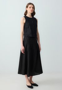Top corto senza maniche nero con una consistenza liscia e una chiusura a bottoni, abbinato a una lunga gonna nera plissettata, caratterizzata da un design fluido.