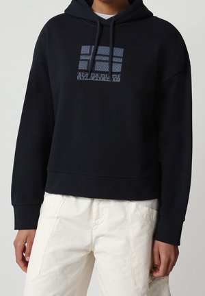 Personne portant un sweat à capuche noir avec un motif rectangulaire bleu à motifs sur la poitrine et un pantalon blanc cassé, debout devant un fond uni.