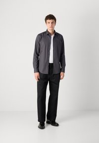 Grünes Langarmshirt mit Knopfleiste, kombiniert mit lockeren schwarzen Hosen und schwarzen Schuhen, vor einem neutralen Hintergrund.