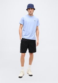 Νεαρός άνδρας που φοράει ανοιχτό μπλε μπλουζάκι, μαύρο σόρτς με ρίγες στο πλάι, λευκά αθλητικά παπούτσια, κάλτσες και μπλε σκουφάκι τύπου κάπα, στέκεται με τα χέρια στις τσέπες.