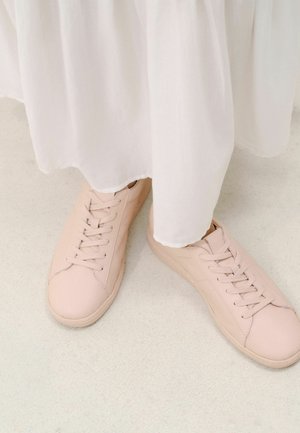 Persoon draagt lichtroze vetersneakers en een witte vloeiende rok, staand op een lichte vloer.