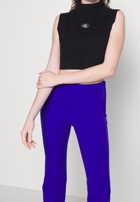 Czarny prążkowany krótki top z wysokim kołnierzem i krótkimi rękawami, z naszywką z logo na przodzie. Noszony z jasnoniebieskimi dopasowanymi spodniami.