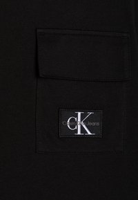 Black cotton shirt featuring a chest pocket, with a white "CK" logo patch and "Calvin Klein Jeans" text beneath, on a smooth texture.