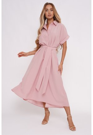 Kobieta o długich blond włosach, ubrana w zwiewną, różową sukienkę midi z paskiem i beżowe sandały na obcasie, stojąca na jasnym jednolitym tle.